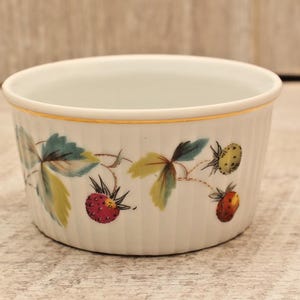 May include: A white porcelain ramekin featuring a delicate fluted exterior and a gold-trimmed rim. The side is decorated with a botanical pattern of green leaves and small red and yellow strawberries, showcasing a classic, clean design.