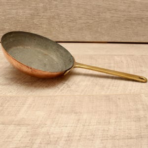 Könnte beinhalten: Eine runde Kupferpfanne mit einer metallischen Innenbeschichtung und einem langen, schlanken Messinggriff, der mit einer kleinen Metallniete befestigt ist. Die Außenseite der Pfanne hat ein warmes, poliertes Kupferfinish, während der Griff glatt und goldfarben ist.