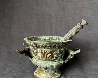 May include: An antique bronze mortar and pestle with ornate detailing. The mortar is shaped like a vase with two handles and the pestle has a rounded end.