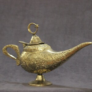 May include: A gold genie lamp with intricate engraved designs and a crescent moon finial. The lamp has a spout and a handle.