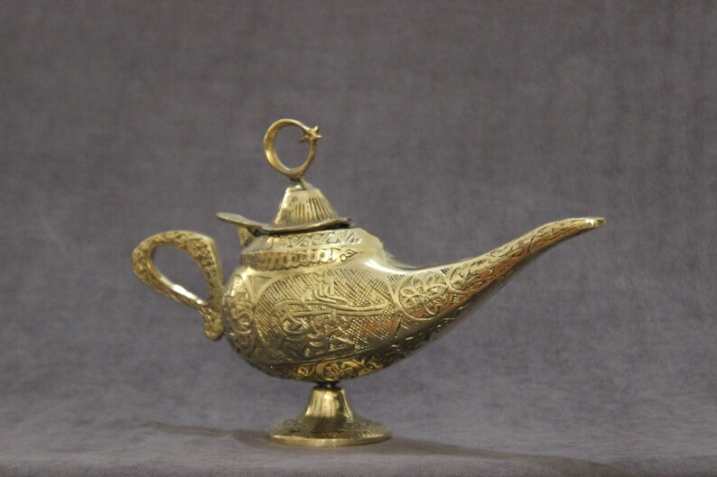 May include: A gold genie lamp with intricate engraved designs and a crescent moon finial. The lamp has a spout and a handle.