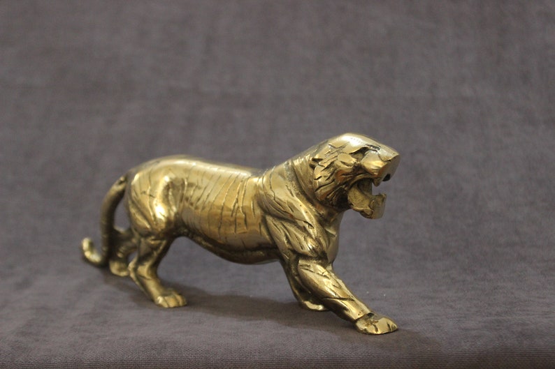 May include: A gold metal tiger figurine with a detailed, textured surface. The tiger is standing on all fours with its mouth open in a roar.