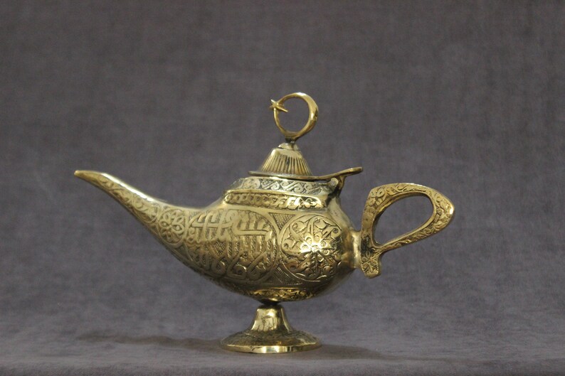 May include: A gold genie lamp with intricate engraved designs. The lamp has a spout, a handle, and a lid with a crescent moon finial.
