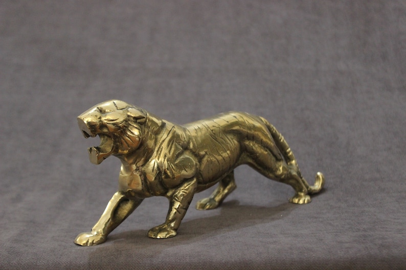 May include: A gold-colored metal figurine of a tiger in a snarling pose. The tiger is depicted with detailed features and a realistic appearance.