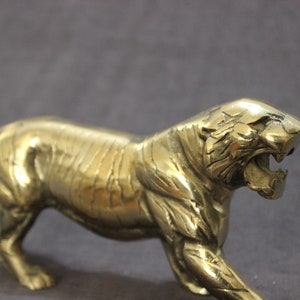 May include: A gold-colored metal figurine of a tiger in a crouching position. The tiger has a detailed face with a wide open mouth and sharp teeth.