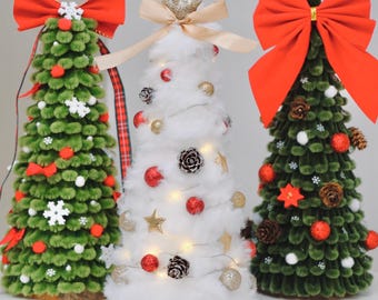 May include: Three cone-shaped decorative trees. The left tree is green with red bows, white snowflakes, and red pom-poms. The center tree is white, fluffy, and adorned with gold and red ornaments, pinecones, and warm string lights. The right tree is green with a large red bow, pinecones, and red spheres.