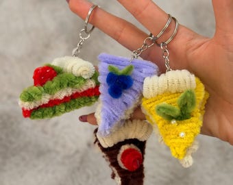 May include: Four slice-shaped keychains made of fuzzy pipe cleaners. One is green with red layers and a red top, one is light purple with blue berries, one is yellow with green leaves and white beads, and one is brown with a red circular top.