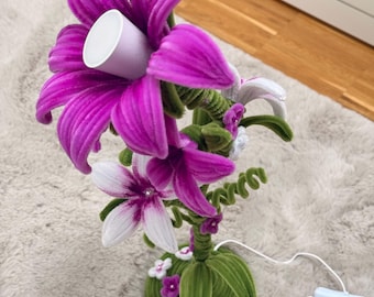 May include: A decorative lamp shaped like a bouquet of flowers with vibrant purple and white petals. The stems are green and textured, rising from a circular green base adorned with small white and purple blossoms. A white power cord is attached.