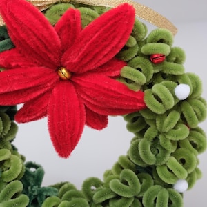 Könnte beinhalten: Ein runder Kranz aus grünen Pfeifenreinigern mit einer großen, samtig roten Weihnachtssternblüte oben, verziert mit kleinen roten Glöckchen, weißen Bommeln und einer goldenen Bandschlaufe zum Aufhängen.