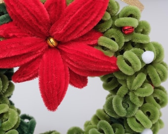 May include: A circular wreath made of green pipe cleaners featuring a large red velvet-textured poinsettia flower at the top, accented with small red jingle bells, white pom-poms, and a gold ribbon loop for hanging.