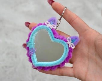 May include: A heart-shaped mirror keychain featuring a fuzzy purple border with light blue trim. The mirror is accented with two small white beaded bows, a translucent blue star charm, and a small purple bear charm on the edges.