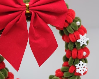 May include: A circular wreath made of alternating red and olive green pipe cleaners, featuring a large red fabric bow at the top. The wreath is decorated with small white plastic snowflake cutouts and tiny white faux pearl beads throughout.