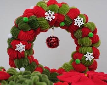 May include: A circular wreath crafted from red and green pipe cleaners, decorated with small white snowflake cutouts, tiny green pom-poms, and a single red jingle bell hanging from the center. A gold string loop is attached to the top.