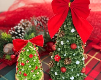 May include: Two cone-shaped Christmas tree decorations made of green textured material. The smaller tree sits on a wood slice base with a red bow and ribbon. The larger tree features a large red bow, pinecones, red glitter balls, and white pom-poms.