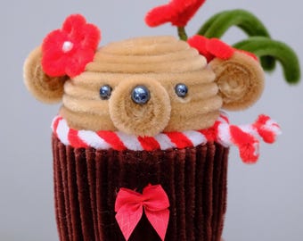 May include: A handcrafted cupcake-shaped figure made from brown and tan pipe cleaners. The top features a bear-like face with dark bead eyes, red flower accents, and a red and white striped scarf. The base is dark brown with a small red bow.