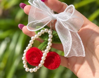 May include: A keychain featuring two red fuzzy circular charms attached to a green stem, accented by a string of white faux pearls and a large, translucent white organza ribbon bow tied at the top, all connected by a silver metal ring.