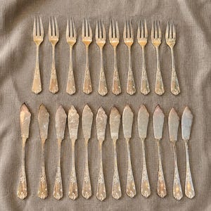 Pode incluir: Uma variedade de talheres de prata vintage, incluindo garfos e facas de manteiga, dispostos sobre um tecido de tom neutro. Os talheres apresentam detalhes ornamentais nas alças, e os dentes dos garfos são visíveis. As facas de manteiga têm uma lâmina plana e arredondada.