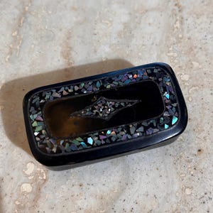 May include: A rectangular black box with a decorative inlay of iridescent pieces around the edges. The center features a stylized diamond shape also inlaid with iridescent pieces.