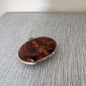 May include: An oval-shaped compact case featuring a polished, mottled brown and amber tortoiseshell-style inlay set within a smooth, metallic silver-toned frame. The lid is slightly domed, showcasing a rich, variegated pattern of dark and light tones.