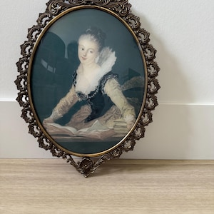 May include: An oval-shaped framed portrait of a woman in a white lace collar and a dark dress, holding an open book. The frame is ornate and gold-coloured.