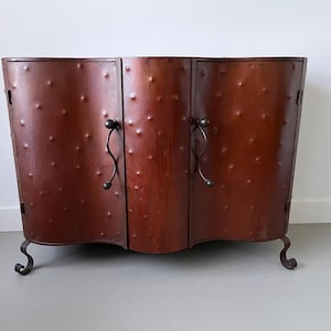 Puede incluir: Un mueble de tres puertas con acabado marrón rojizo oscuro. Las puertas del mueble tienen una superficie texturizada con pequeños bultos espaciados uniformemente. Cuenta con tiradores curvos de hierro forjado y patas decorativas. El mueble mide aproximadamente 122 cm de ancho.