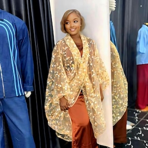 Könnte beinhalten: Eine Frau trägt einen durchsichtigen, hellgelben Kimono mit floralem Muster über einem rostfarbenen Kleid. Der Kimono hat ein fließendes Design. Der Hintergrund umfasst eine Schaufensterpuppe und Kleiderauslagen.