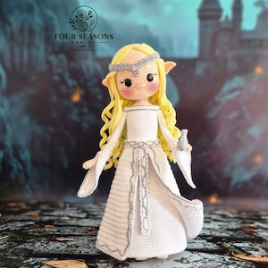 May include: A crocheted doll with long wavy yellow hair, pointed ears, and rosy cheeks. The doll wears a white floor-length gown with silver trim, a matching silver headband, and holds a small grey object. Text reads FOUR SEASONS SPRING.