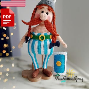 May include: Crochet amigurumi pattern for a Viking warrior doll with a blue and white striped shirt, a brown beard, and a grey helmet. The doll is holding a blue shield with a yellow circle in the center. The pattern is available as a PDF download.
