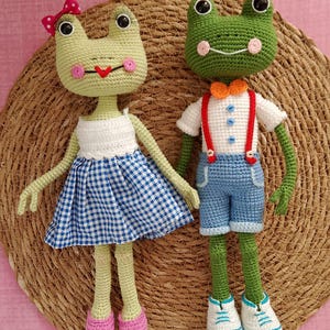 May include: Two crocheted frog plushies, one wearing a blue and white gingham dress with a pink bow and the other wearing a white shirt, blue overalls, and a bow tie. The plushies are sitting on a woven straw mat.