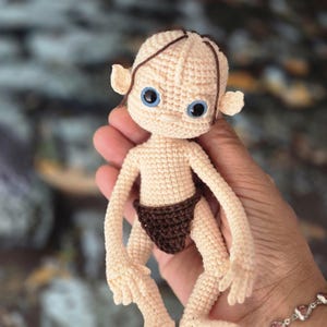Può includere: Una piccola bambola lavorata all'uncinetto a mano con un incarnato beige pallido, grandi occhi blu in plastica e sottili fili di lana marrone come capelli. La figura indossa un perizoma all'uncinetto marrone scuro e ha arti lunghi.
