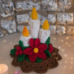 Puede incluir: Centro de mesa de velas de ganchillo con tres velas blancas rematadas con llamas amarillas. Una flor roja grande y flores rojas más pequeñas con hojas verdes están en el centro, todo sobre una base de ganchillo marrón. El texto "Four.seasons_spring" es visible.