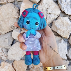 Puede incluir: Una muñeca de crochet con pelo azul, detalles rosas y un vestido de lunares morados y blancos. La muñeca tiene las manos juntas frente a su pecho. La muñeca está sentada en una mano con una pulsera en la muñeca.