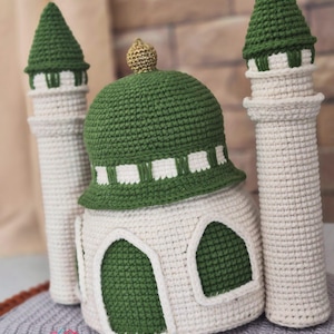 May include: A handmade crochet mosque with a green dome and minarets. The mosque is white with green accents, including the dome, arched windows, and the tops of the minarets. A gold-colored finial tops the dome. The base of the mosque is white with green arched windows.