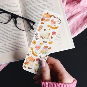 Puede incluir: Un marcap&aacute;ginas blanco con ilustraciones de tem&aacute;tica oto&ntilde;al, que incluyen calabazas, libros, hojas y una bufanda. El marcap&aacute;ginas se sostiene frente a un libro abierto y un su&eacute;ter rosa. La palabra "books" est&aacute; escrita en el marcap&aacute;ginas.