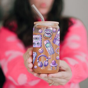 May include: A clear glass tumbler with a wooden lid and a stainless steel straw, decorated with purple and white retro-themed stickers. The stickers include a cassette player, a cell phone, headphones, and smiley faces. The tumbler is filled with a brown liquid.