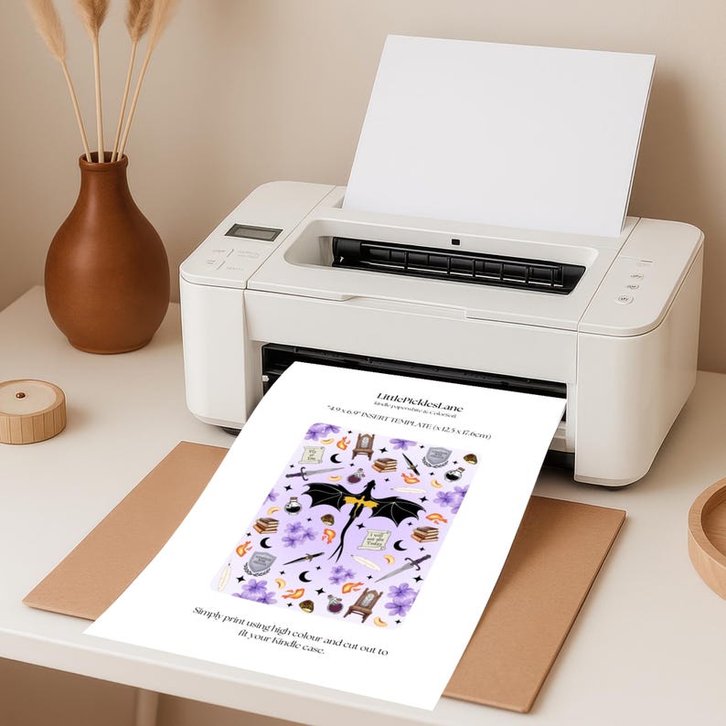 Op de afbeelding: Een witte printer die een kleurrijke inlegsjabloon met een drakenontwerp afdrukt. De sjabloon bevat zwaarden, boeken en bloemen op een lavendelkleurige achtergrond. De tekst op de sjabloon luidt "LittlePicklesLane" en "Simply print using high colour and cut out to fit your kindle case."