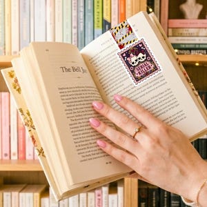 Puede incluir: Un marcap&aacute;ginas con un dise&ntilde;o de sello rectangular con borde morado, dos calaveras blancas con lazos rosas y el texto Serial Killer en una fuente decorativa, rematado con una secci&oacute;n de patr&oacute;n de cinta de precauci&oacute;n amarilla y negra.