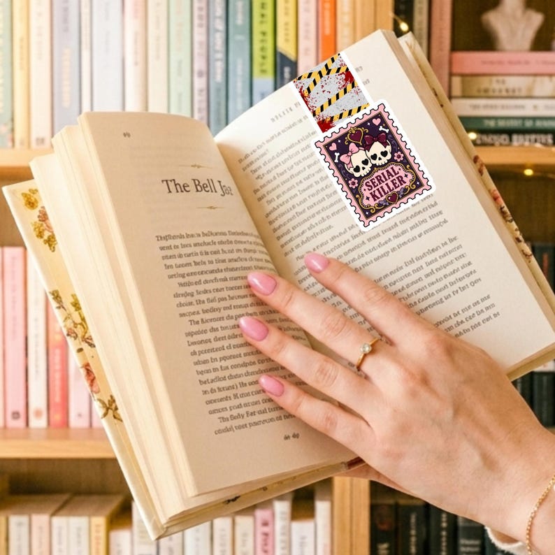 Puede incluir: Un marcap&aacute;ginas con un dise&ntilde;o de sello rectangular con borde morado, dos calaveras blancas con lazos rosas y el texto Serial Killer en una fuente decorativa, rematado con una secci&oacute;n de patr&oacute;n de cinta de precauci&oacute;n amarilla y negra.