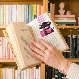 May include: A bookmark featuring a black cow wearing pink glasses and holding a pink book, placed in an open book. The bookmark has a pink and white checkered pattern at the top. The book is open to a page with text.