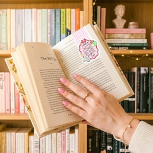 May include: An open book with a pink and white striped bookmark that reads "Sorry I have a little bookish life". The book is held open by a hand with a gold ring and bracelet. Bookshelves are in the background.