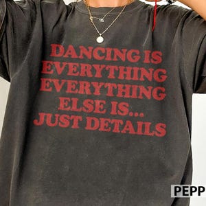 May include: A charcoal gray short-sleeve t-shirt featuring red text that reads DANCING IS EVERYTHING EVERYTHING ELSE IS... JUST DETAILS. The shirt has a crew neckline and a relaxed fit, showcasing a vintage-inspired washed fabric texture.
