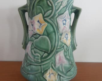 Puede incluir: Un jarrón de cerámica verde con dos asas laterales y un patrón en relieve de flores y enredaderas de gloria de la mañana. Las flores están coloreadas en tonos suaves de rosa, amarillo y azul sobre una superficie esmaltada en verde mate.