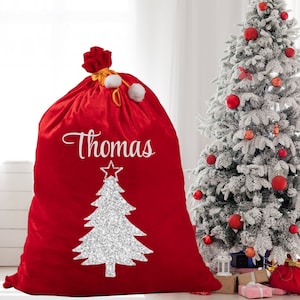 May include: A large red Christmas sack with the name "Thomas" in white script. A silver glitter Christmas tree design is below the name. The sack is tied at the top with a red bow and white pom-poms. A decorated Christmas tree is in the background.