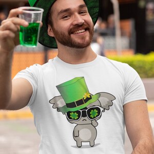 May include: A white t-shirt with a cartoon koala wearing a green leprechaun hat and sunglasses with shamrock designs. The koala is holding a shamrock in its hand.