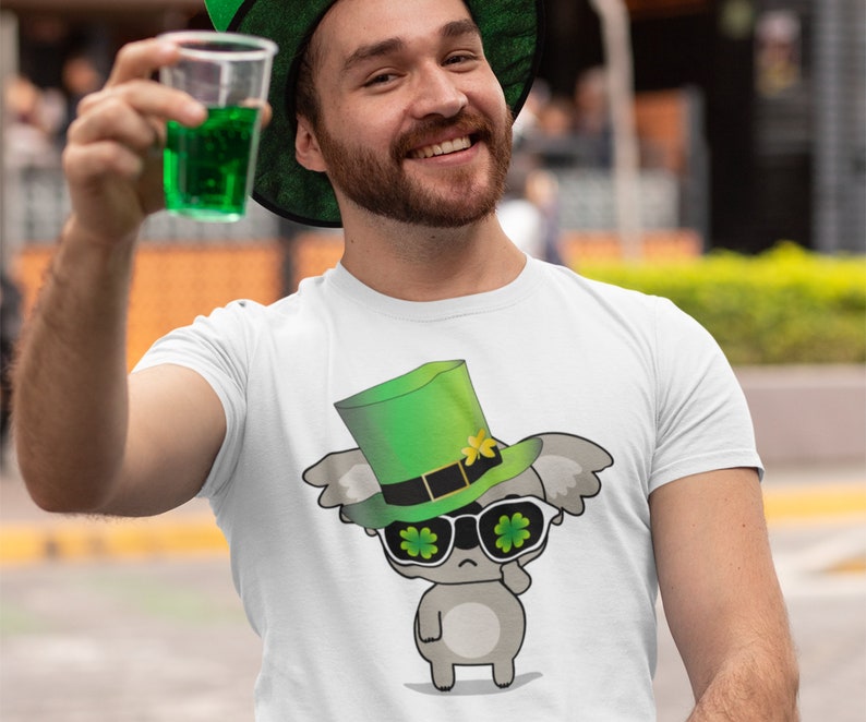 May include: A white t-shirt with a cartoon koala wearing a green leprechaun hat and sunglasses with shamrock designs. The koala is holding a shamrock in its hand.