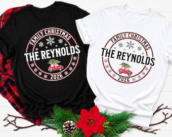 Peut inclure: Un t-shirt noir et un t-shirt blanc, chacun arborant un motif circulaire avec le texte FAMILY CHRISTMAS, THE REYNOLDS et 2026. Le design comprend une voiture rouge transportant un sapin de Noël vert et des flocons de neige blancs.