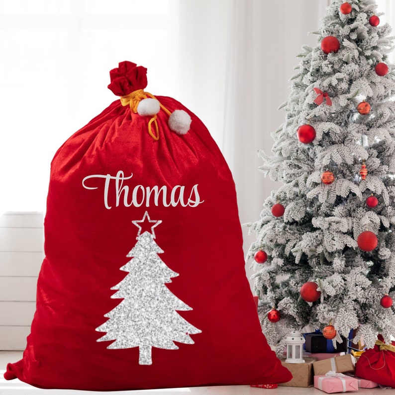 May include: A large red Christmas gift sack with the name "Thomas" in white script. A silver glitter Christmas tree design is below the name. The sack is tied at the top with a red bow and white pom-poms. A decorated Christmas tree is in the background.