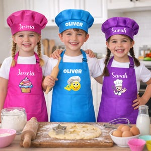 May include: Three children wearing personalised chef hats and aprons in pink, blue, and purple. The pink apron features the name Jesscia and a cupcake graphic. The blue apron displays the name Oliver and a smiley face. The purple apron shows the name Sarah and a chef illustration.