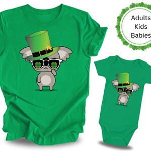 May include: Green t-shirt and bodysuit with a cartoon koala wearing a green leprechaun hat and sunglasses with shamrocks. The text "Adults Kids Babies" is on a green shamrock background.