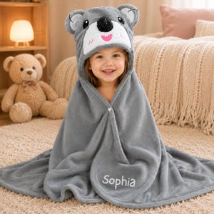 May include: A grey hooded towel featuring a koala bear design on the hood. The towel has a button fastening and the name "Sophia" embroidered on the front. A child is smiling, wrapped in the towel.